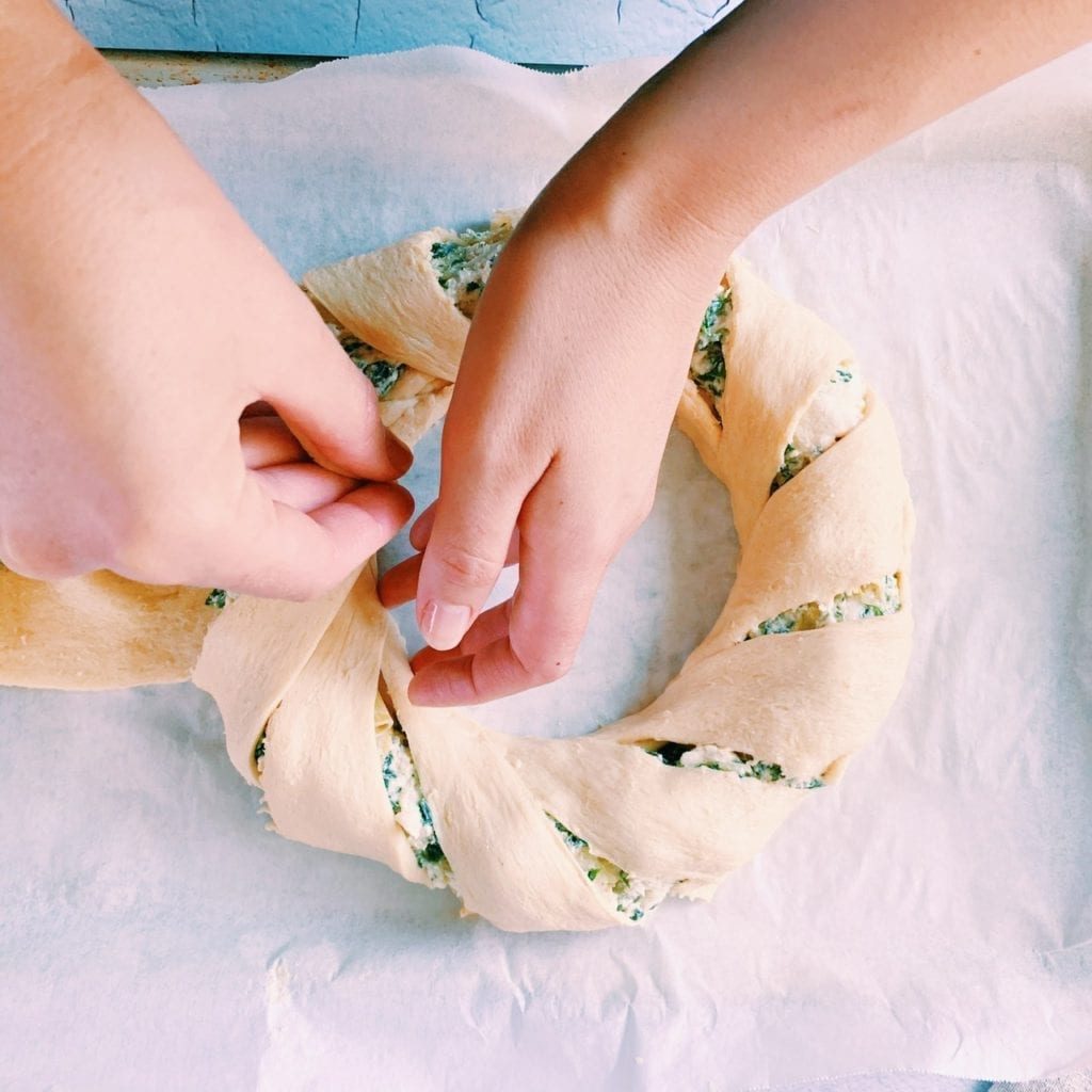 Two hands pulling the tip of a crescent roll triangle over spinach and artichoke dip ring and tucking it in the center