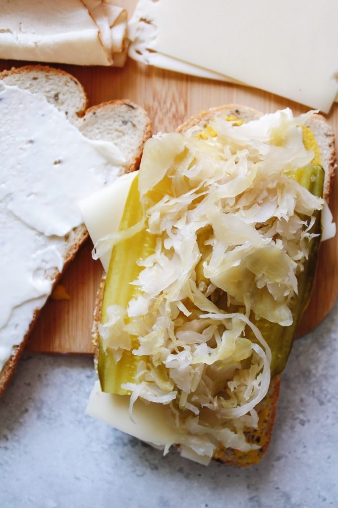 sandwiches being assembled with pickles, saurkraut, turkey and mustard on a cutting board