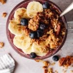 cherry berry oat milk smoothie in a clear glass mug topped with granola, bananas and blueberries
