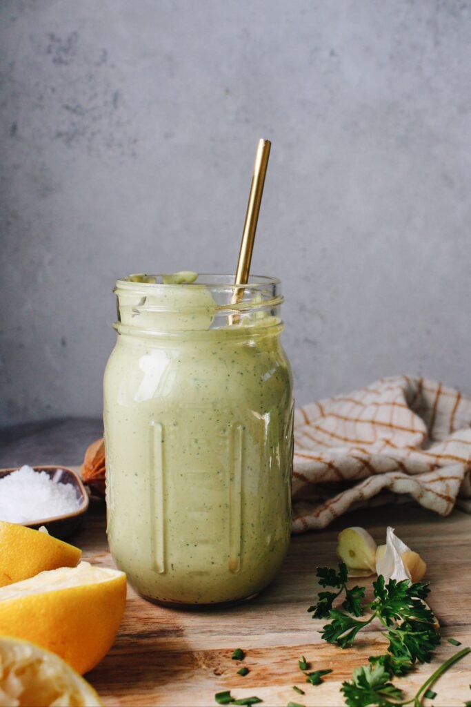 lemon herb tahini sauce in a jar surrounded by lemon, parsley, garlic