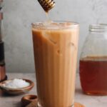 an iced honey cinnamon latte in a clear glass with a honey dipper drizzling honey