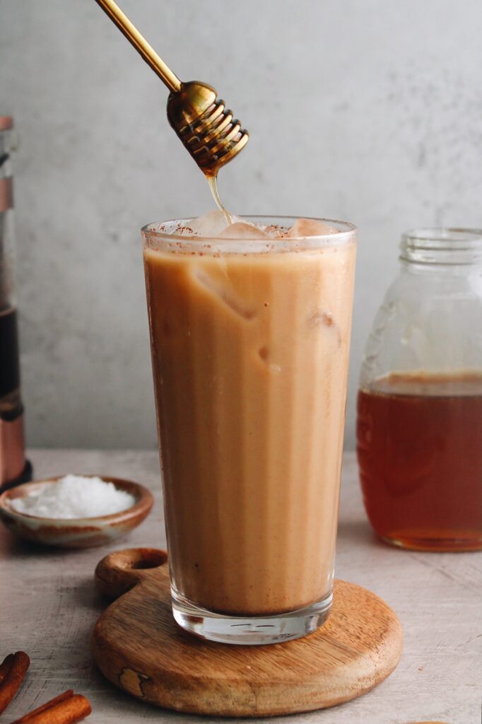 an iced honey cinnamon latte in a clear glass with a honey dipper drizzling honey