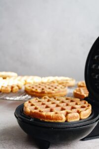 cooked waffle sitting in waffle iron with more waffles on cooling rack behind it