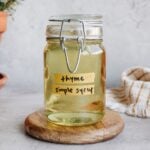thyme simple syrup in a clear glass jar sitting on a white coaster