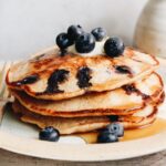 stack of blueberry lemon pancakes on a white plate topped with blueberries and syrup