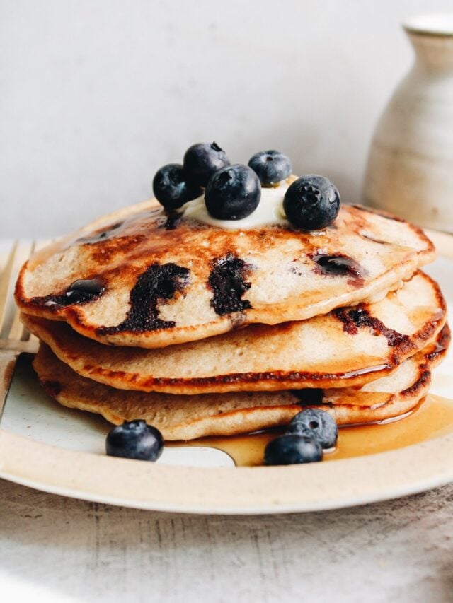 stack of blueberry lemon pancakes on a white plate topped with blueberries and syrup