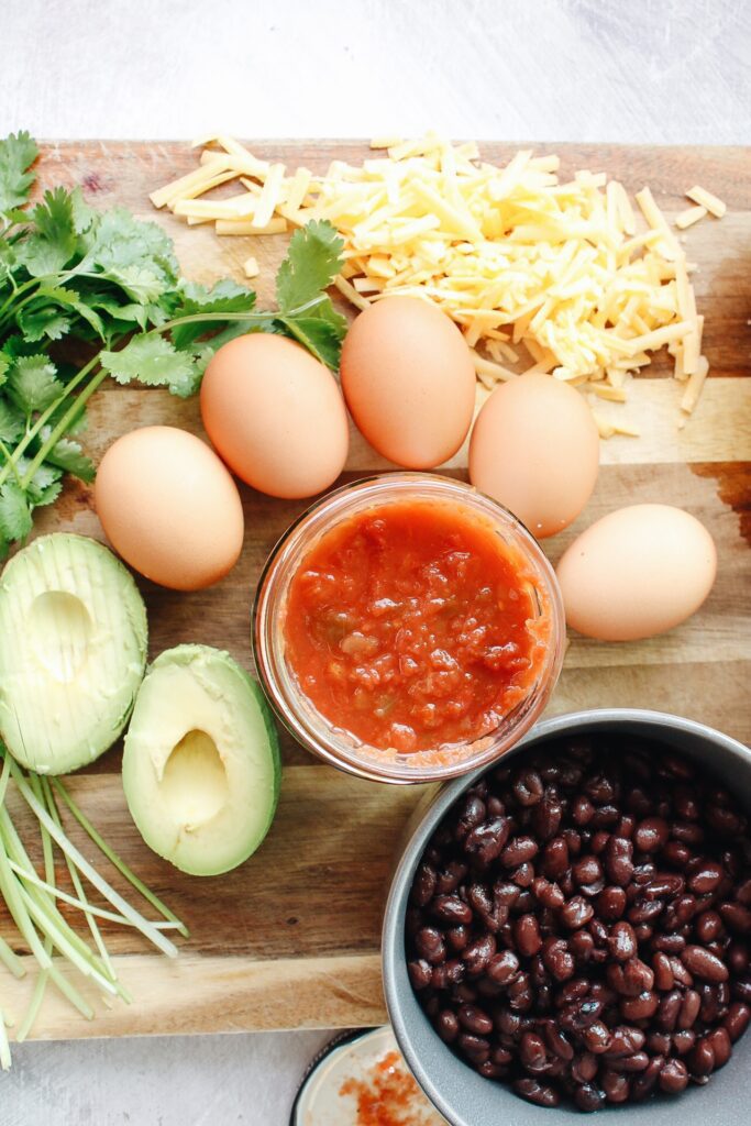 ingredients for southwest breakfast bowl on a cutting board: black beans, avocado, eggs, salsa, cheddar cheese and cilantro