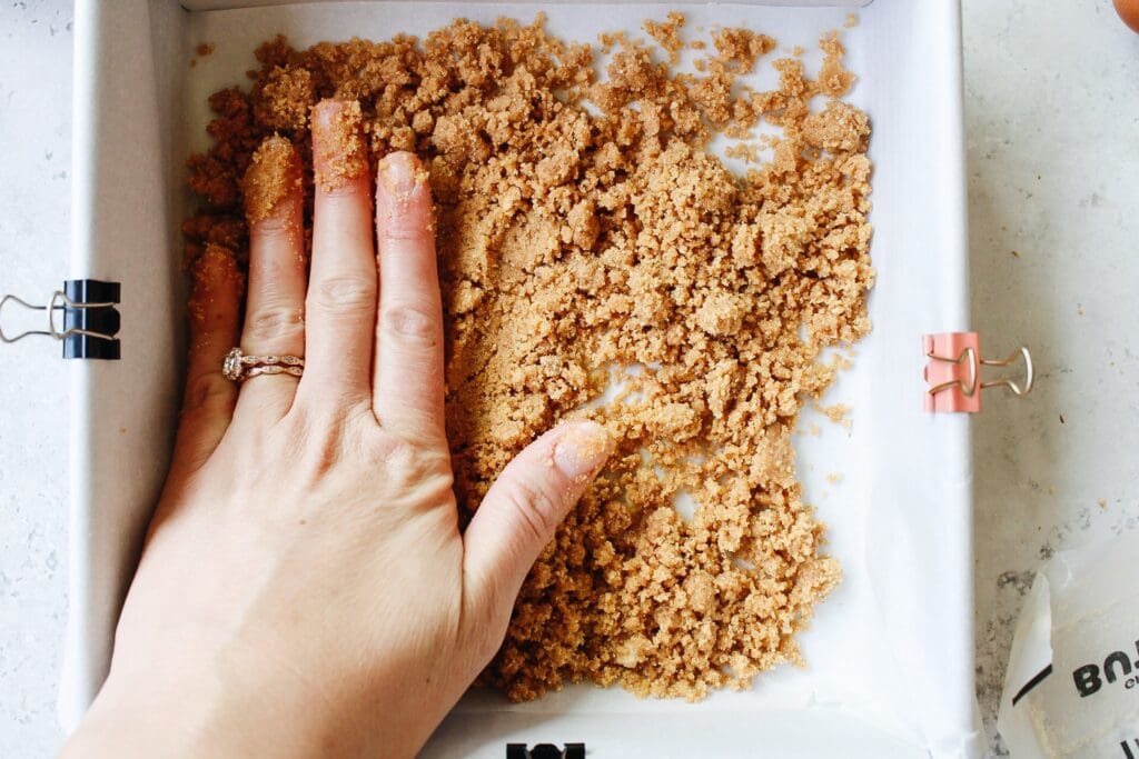 graham cracker crust mixture being pressed into the bottom of a baking pan