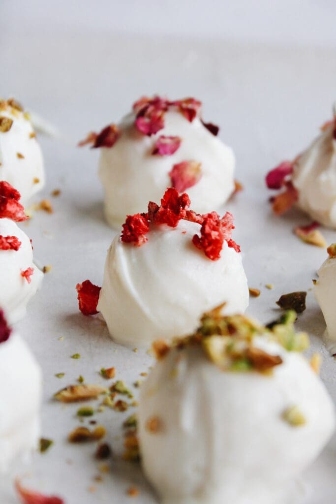 rose white chocolate truffles on a baking sheet topped with chopped pistachio, strawberries and rose petals. 