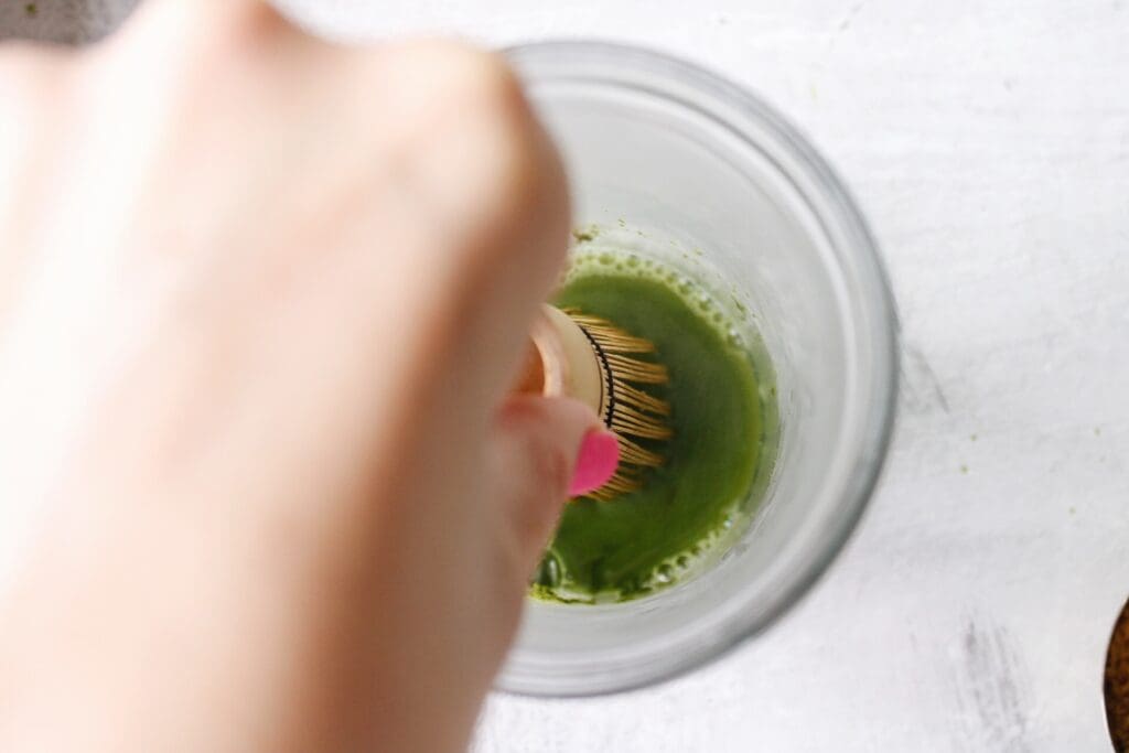 matcha and hot water being mixed with a matcha whisk in a glass