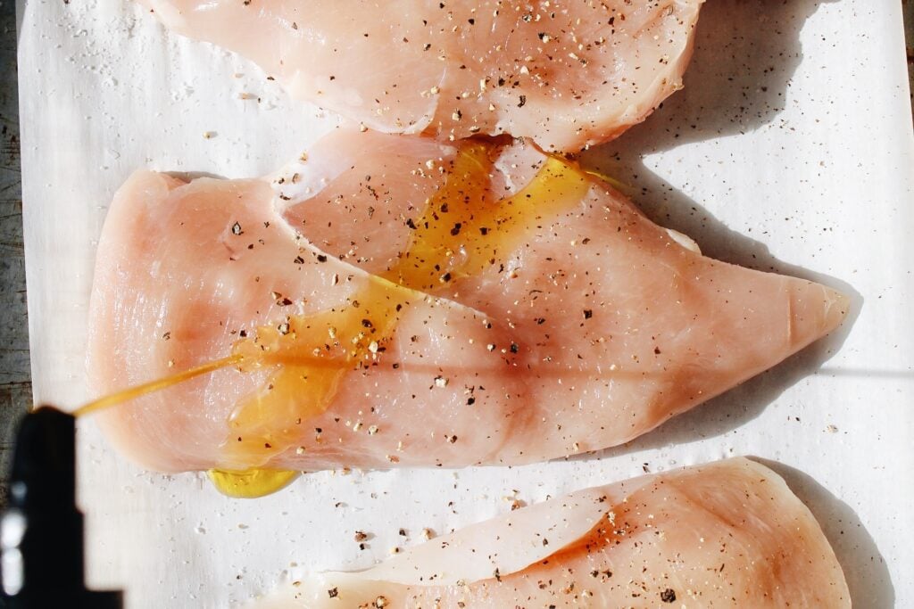 olive oil being drizzled on chicken with black pepper